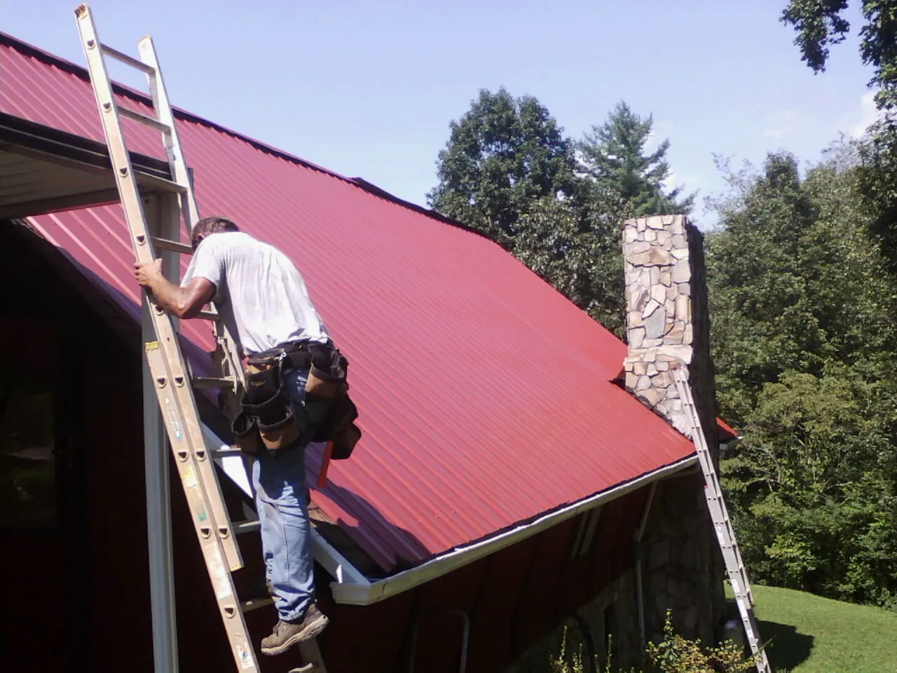Roofer on a ladder beside a red metal roof and stone chimney — Soffit & Fascia Repair by Roofing Brothers in Ransomville