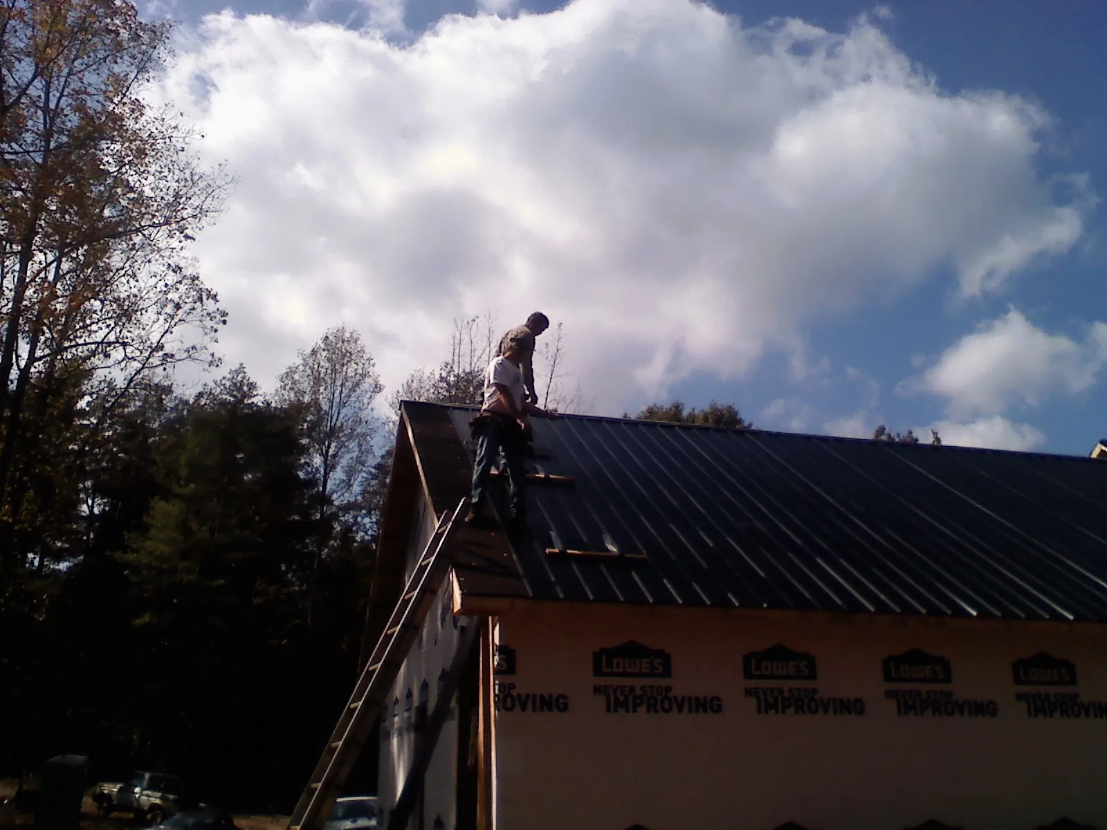 Roofer fastening dark metal panels high against a cloudy sky, from a Roof Inspection job in Ransomville